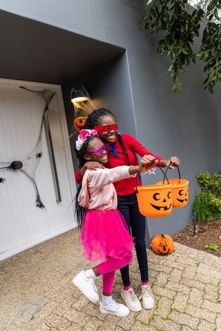 African american sisters trick-or-treating in bright costumes holding pumpkin buckets