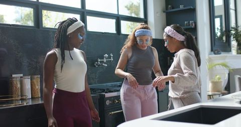 African american friends pampering with facial masks and headbands in sunlit modern kitchen