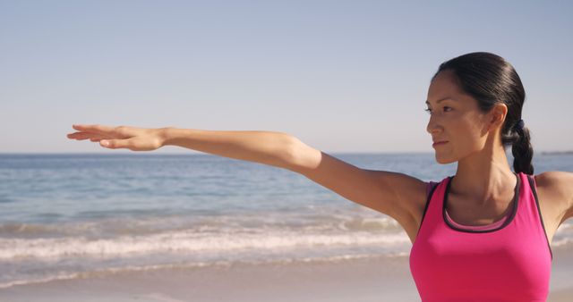 Woman with dark hair and pink top practicing yoga on beach in morning, reaching out arms. Calm sea in background and clear sky add peaceful and serene atmosphere, ideal for themes related to wellness, fitness, outdoor activities. Suitable for articles, blogs, or promotions about health and fitness, mental well-being, yoga practices.