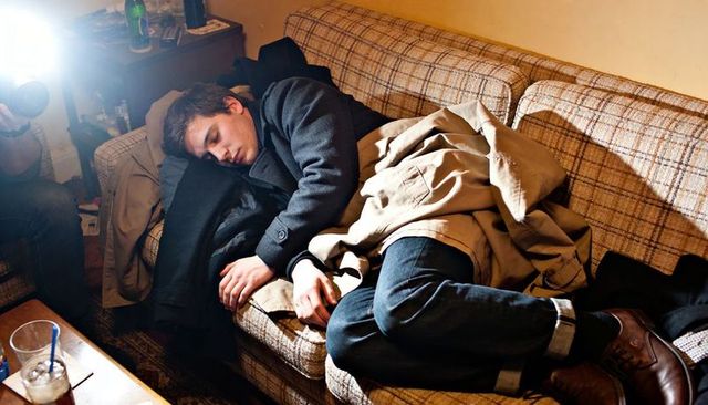 Young man sleeping on plaid sofa wearing trench coat and jeans in messy living room