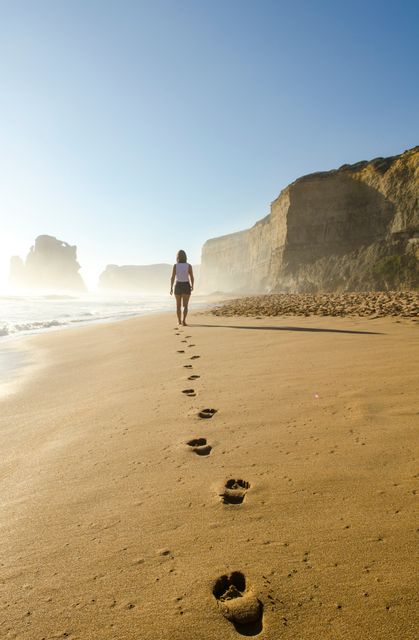 Woman walking by herself on a sandy beach with footprints in sand and large cliffs in background. Remote coastal location offers serene and tranquil vibe, perfect for nature-themed travel or lifestyle blogs. Ideal for content about solo adventures, serene escapes, or outdoor activities.
