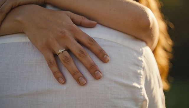 Sunlit couple embracing while cradling pregnant belly, hand showing gold engagement ring