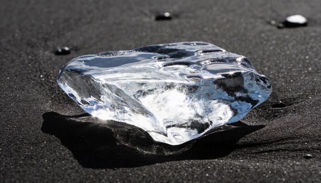 Glittering ice chunk sitting on black sand beach reflecting sunlight and wet pebbles