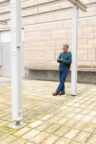 Senior man leaning on pergola post checking smartphone in urban courtyard