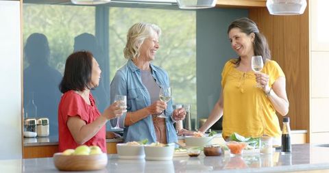 Mature women laughing and sharing wine while preparing healthy meal at modern kitchen island