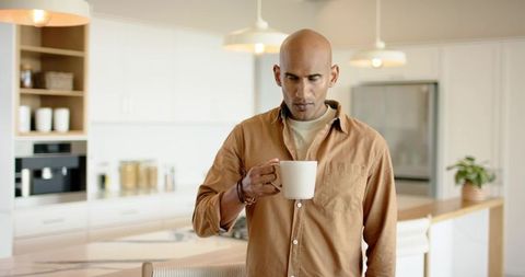 Mature bald man sipping coffee at minimalist sunlit kitchen island in casual tan shirt