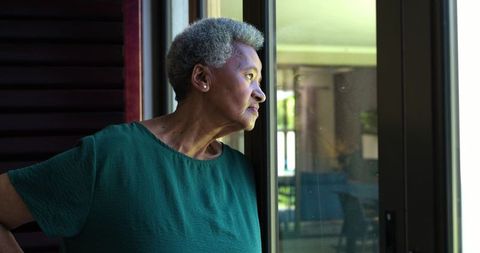 Senior african american woman gazing through sliding glass door into sunlit patio