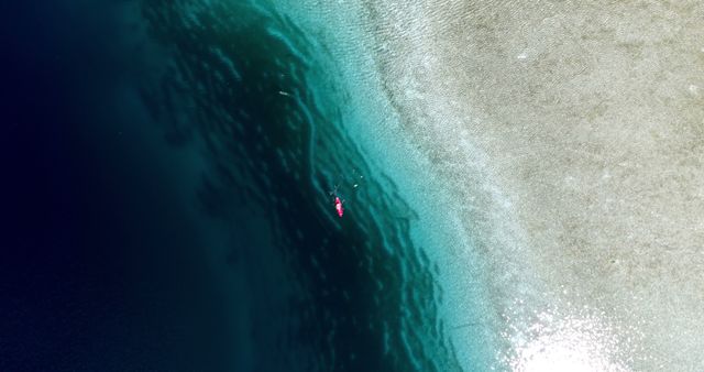 Aerial view of a person floating on a clear blue sea near a sandy beach, with copy space. The contrast between the deep blue water and the light sand creates a serene and inviting landscape for relaxation or water activities.