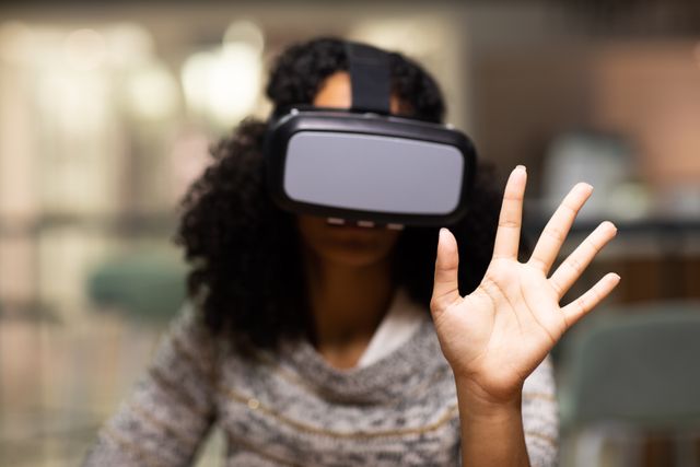 Front view of a biracial professional woman working late in a modern office, sitting at a desk wearing a VR headset, with one hand raised in front of her