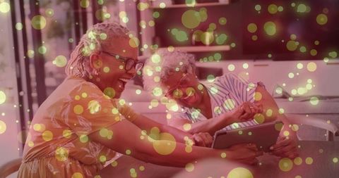 Laughing senior women sharing tablet joy in cozy lounge with golden bokeh light overlay