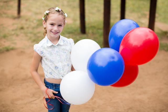 A young girl is holding red, white, and blue balloons while smiling in a park. She is wearing a floral shirt and has a flower headband in her hair. This image can be used for themes related to childhood, outdoor activities, celebrations, and festive events.