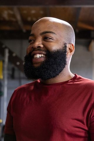 Smiling african american man standing in garage gym wearing maroon t-shirt, looking left
