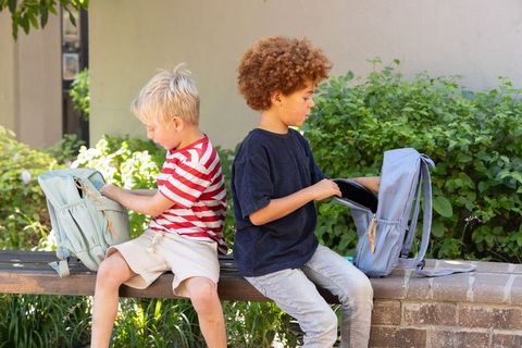 Two boys searching backpacks on bench in sunny school courtyard showing friendship