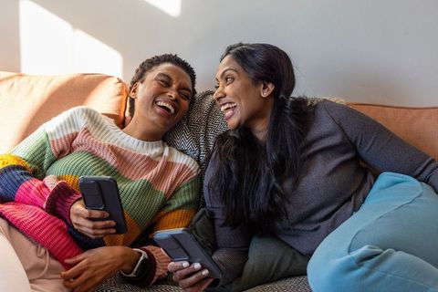 Friends sharing laughs on sofa while using smartphones in sunlit living room
