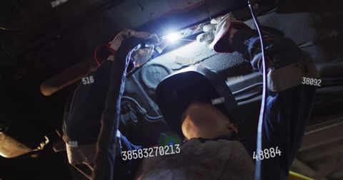 Female welder welding under vehicle holding torch wearing helmet gloves in garage workshop