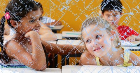 Smiling schoolgirls leaning together at desks with math formulas overlaying glass