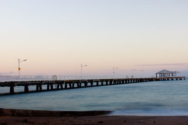 The image captures a long wooden pier extending into calm blue ocean waters under a sunset sky. The horizon line is visible with soft evening light creating a tranquil and serene atmosphere. Perfect for illustrating concepts of relaxation, tranquility, and coastal living. Ideal for use in travel blogs, inspirational posters, websites related to beach life, and social media content.