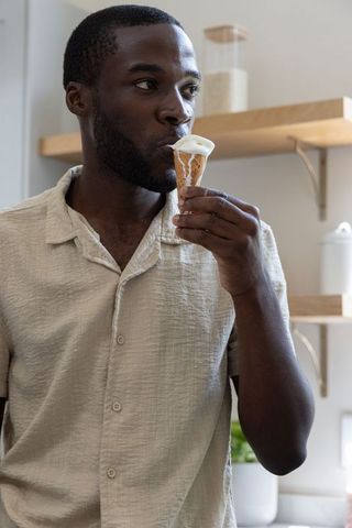 African american man enjoying melting ice cream cone in bright kitchen licking waffle cone