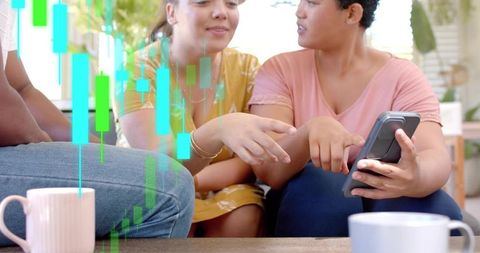 Young women sharing smartphone and analyzing investment charts in casual living room