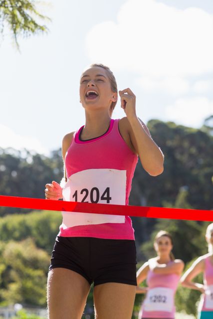 Cheerful female athlete crossing the finish line in a park race, celebrating her victory. Perfect for use in advertisements for sportswear, fitness events, marathon promotions, and inspirational content about determination and success.
