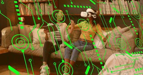 Two women using vr headset and tablet with futuristic augmented interface on cozy living room sofa