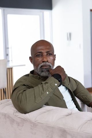 Mature african man sitting on beige tufted sofa resting chin, contemplating by window