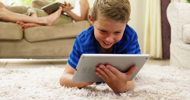 Young boy lying on carpet and using tablet, smiling happily. Scene suggests a modern home environment with focus on digital learning and enjoyment. Ideal for educational resources, technology integration, child-friendly gadget promotions, family lifestyle, and home decor themes.