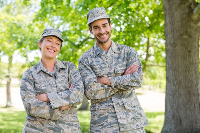 This image shows a smiling military couple standing with arms crossed in a park on a sunny day. Both individuals are wearing military uniforms, indicating their service. The background features lush green trees and a bright, sunny environment. This image can be used for themes related to military life, camaraderie, teamwork, confidence, and outdoor activities.