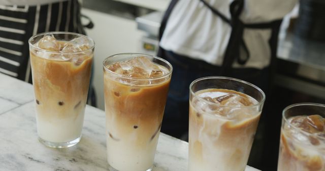 Four fresh iced lattes sit on a marble counter, each glass filled with layers of coffee and milk, while baristas in striped and navy aprons are preparing more drinks in the background. Perfect for advertisements or blog posts about coffee culture, beverage recipes, and cafe promotions.