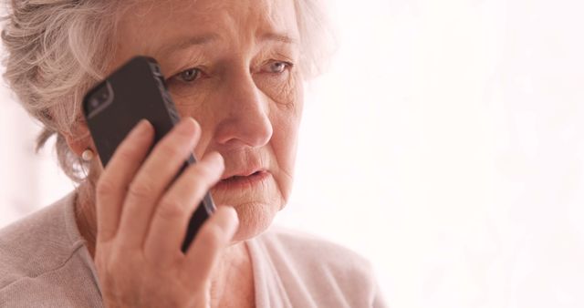 Elderly woman with grey hair talking on mobile phone at home, looking concerned. Captures senior lifestyle, communication technology usage, and emotional moments. Useful for topics about elderly care, mental health, and family support.