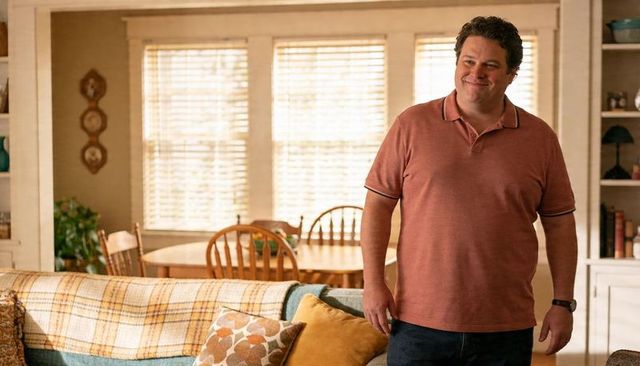 Smiling man standing in cozy living room wearing rust polo and dark jeans, plaid throw