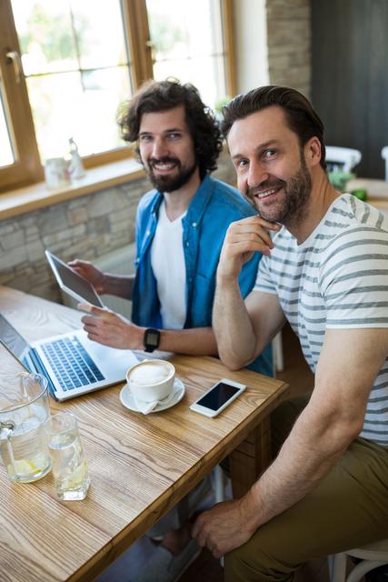 Two men are sitting at a wooden table in a coffee shop, engaging with a digital tablet and a laptop. They are smiling and appear to be enjoying their time together. This image is ideal for illustrating concepts of teamwork, remote work, modern lifestyle, and technology in a casual setting. It can be used in articles, blogs, or advertisements related to business, technology, or social interactions.