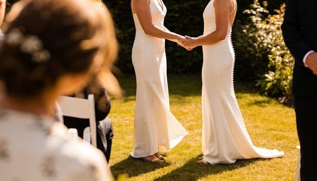 Two brides holding hands during sunlit garden wedding ceremony wearing elegant gowns
