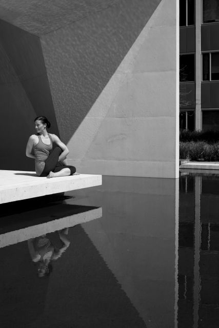 Woman practicing seated yoga pose in outdoor space near modern architectural structure. Reflection on water adds serene atmosphere. Ideal for promoting wellness, exercise, meditation, and urban lifestyle topics.