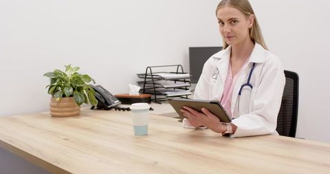 Female doctor using tablet at minimalist clinic desk with stethoscope and coffee cup