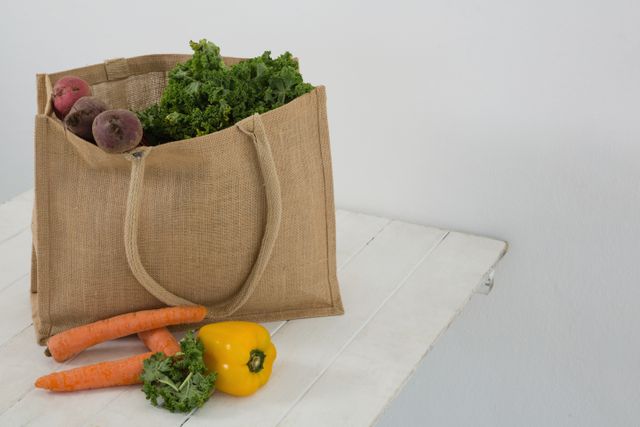 Eco-friendly grocery bag filled with fresh vegetables including carrots, bell pepper, and kale on a white table. Ideal for promoting healthy eating, organic produce, sustainable living, and zero waste lifestyle. Perfect for use in articles, blogs, and advertisements related to nutrition, eco-friendly products, and farm-to-table concepts.