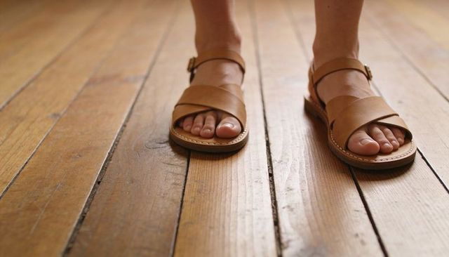 Female feet standing in tan leather sandals on varnished hardwood floor, close-up