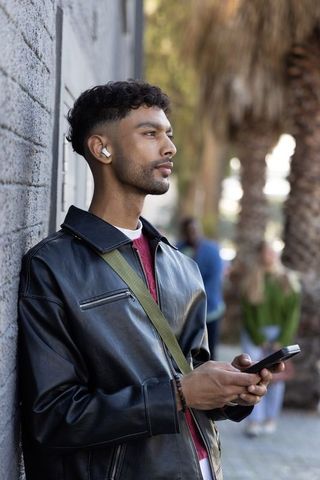 Indian man leaning on brick wall holding smartphone wearing leather jacket and earbuds