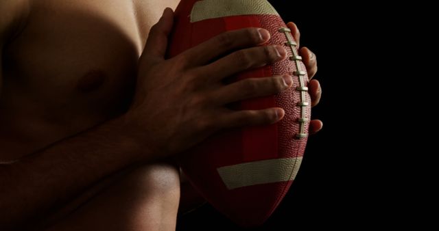 Close-up image of a muscular man gripping a football against a dark background emphasizes dedication, strength, and athleticism. Ideal for use in sports promotion, fitness and training materials, team-based athletic campaigns, motivational posters, and football event advertisements. Conveys themes of preparation, focus, and physical fitness.