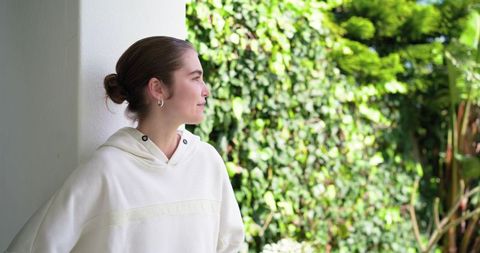 Young woman leaning on white wall gazing at garden in white hoodie
