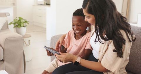 Woman guiding child on tablet while smiling on couch, family bonding and digital learning