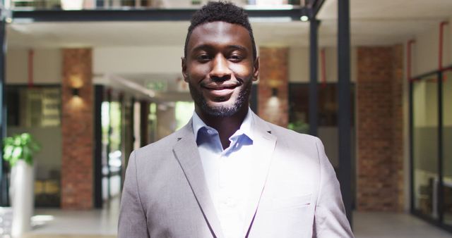 Smiling African American businessman posing in modern office corridor filled with natural light. Wearing a suit, demonstrating professionalism and confidence. Ideal for business, career, corporate culture, diversity, and workplace concepts.