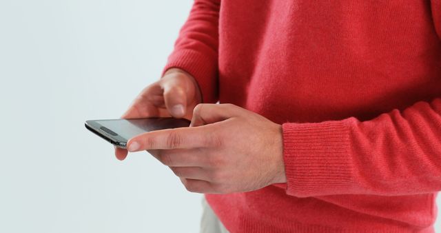 A man wearing a red sweater using a smartphone against a plain white background. He is holding the phone with both hands and appears to be texting or interacting with the screen. The casual attire and modern technology theme make this perfect for illustrating concepts related to everyday communication, tech-savvy lifestyles, or mobile usage in marketing materials, website layouts, and lifestyle blogs.