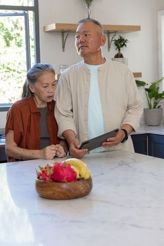 Senior asian couple using tablet in modern bright kitchen with dragon fruit bowl