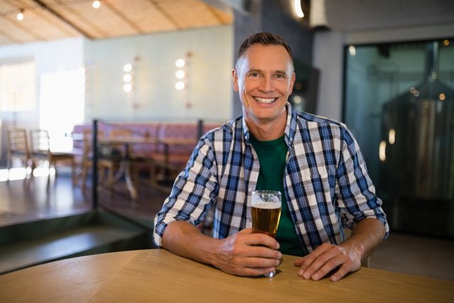 This image features a cheerful man sitting at a counter in a modern bar, enjoying a glass of beer. Ideal for use in advertisements for bars, breweries, or hospitality services. Can also be used in lifestyle blogs, promotional materials for casual dining places, or social media posts focusing on leisure activities.