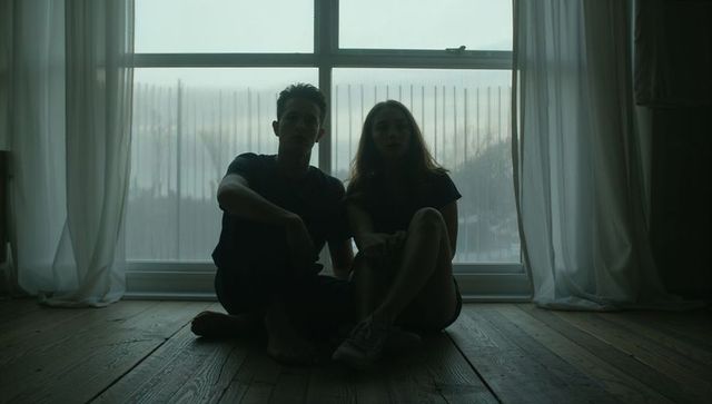 Silhouette couple sitting by window with sheer curtains on wooden floor, moody backlit scene