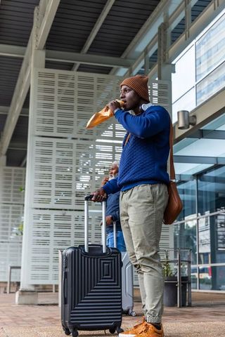 Urban traveler eating sandwich while waiting with suitcase at modern transit terminal