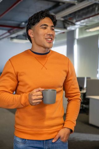 Asian man standing in modern open-plan office enjoying coffee wearing orange sweatshirt