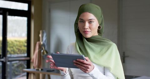 Asian woman wearing green hijab working remotely on tablet in modern home office