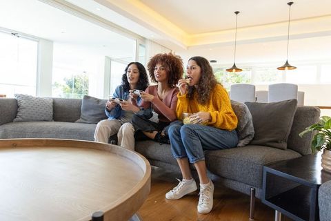 Diverse friends playing console game on sofa, snacking and laughing in modern living room